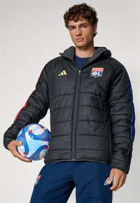 Svart polstret jakke med røde og blå detaljer, glidelåslukking og logo på brystet. Mann som holder en blå og rød mønstrete fotball.