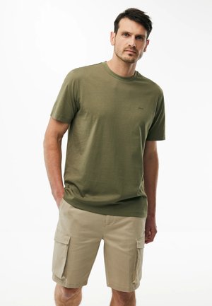 Mann trägt olivgrünes T-Shirt mit kurzen Ärmeln und beige Cargo-Shorts, steht mit einer Hand in der Tasche vor weißem Hintergrund.