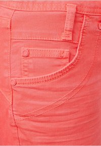 Jeans de denim coral con un ajuste slim. Detalles visibles en los bolsillos y costuras. Textura suave con un ligero brillo. Presenta dos botones en los laterales.