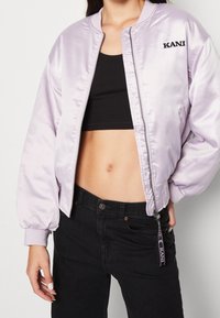 Lila satinkavaj med ribbade manschetter, dragkedja fram och broderad "KANU"-logotyp som accent. Kombineras med en svart kort topp och mörka jeans.