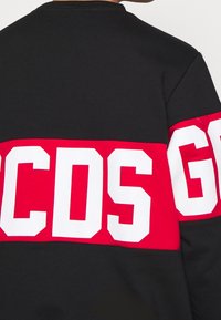 Felpa nera caratterizzata da una ampia fascia rossa decorata con lettere bianche in grassetto che formano la scritta "GCDS." Il collo è rotondo e il tessuto sembra liscio.