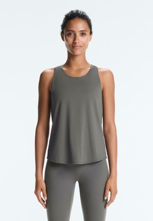 Mladá žena s kučeravými vlasmi oblečená v sivom športovom tielku bez rukávov a zodpovedajúcich legínach, stojaca pred jednoduchým svetlým pozadím.