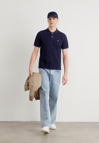 Marinblå polo t-shirt, ljusblå jeans, beige jacka över armen, vita sneakers, mörk keps. Slät tyg med klassisk krage och logodetalj.