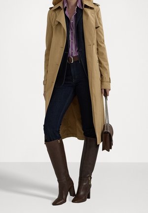Persoon die een beige trenchcoat draagt, een paarse gestreepte blouse, donkere spijkerbroek, bruine kniehoge laarzen en een bruine handtas met kettingriemen vasthoudt.