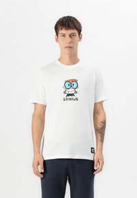 Ung mand med tatovering på venstre underarm iført hvid T-shirt med en tegneseriefigur og teksten "GENIUS", parret med mørke bukser.