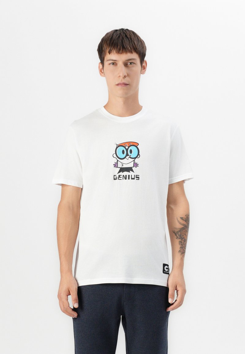 Ung mand med tatovering på venstre underarm iført hvid T-shirt med en tegneseriefigur og teksten "GENIUS", parret med mørke bukser.
