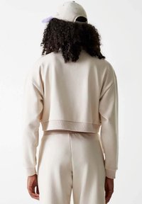 Felpa cropped beige chiaro con polsini e orlo a coste, vista da dietro, abbinata a pantaloni coordinati, texture liscia, design minimale.