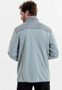 Lyseblå fleecejakke med en grå skulderpanel, der har krave og tekstureret stof. Designet til komfort og varme.