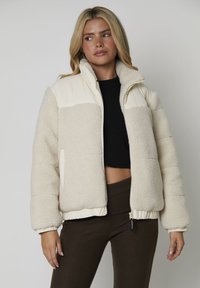 Mujer con cabello largo y rubio, vistiendo una chaqueta de sherpa color crema sobre un top corto negro y pantalones marrón oscuro, frente a un fondo gris liso.