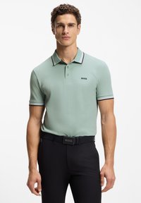 Mintgrøn poloshirt med en ribbet krave med sorte og hvide detaljer, kombineret med sorte bukser og et sort bælte med mærkelogo.
