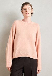 Pull en tricot rose surdimensionné avec un ourlet et des poignets côtelés ; présente un col rond et un tissu doux et texturé. Associé à un pantalon noir.