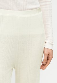 Ριμπ πλεκτό παντελόνι σε κρεμ χρώμα με ελαστική μέση, με ομαλή υφή και διακριτικό κάθετο σχέδιο, εμφανιζόμενο κοντά σε ένα χέρι.