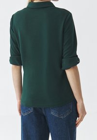 Ciemnozielona koszulka polo z podwiniętymi rękawami, wykonana z miękkiego materiału, z kołnierzykiem i prostym wykończeniem, noszona z niebieskimi dżinsami.