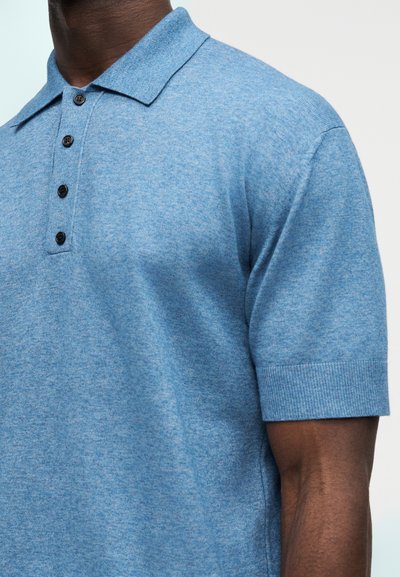 Κοντινό πλάνο ενός ατόμου που φοράει ανοιχτό μπλε κοντομάνικο πλεκτό πόλο με γιακά και τέσσερα μαύρα κουμπιά.