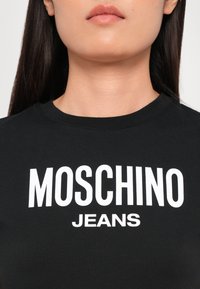 Juodas medvilnės marškinėliai su ryškiai balta tekstu "MOSCHINO JEANS" priekyje. Apvalus iškirpimas, raštuota apdaila, prigludęs dizainas.