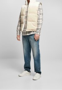 Beige, stoppad puffer-väst över en rutmönstrad skjorta, kombinerad med ljus tvättade jeans och vita sneakers, med en avslappnad passform och en avslappnad stil.