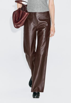 Vrouw in hoge, donkerbruin leren broeken, grijs topje, met een bijpassende leren handtas en zwarte gestikte enkellaarsjes.