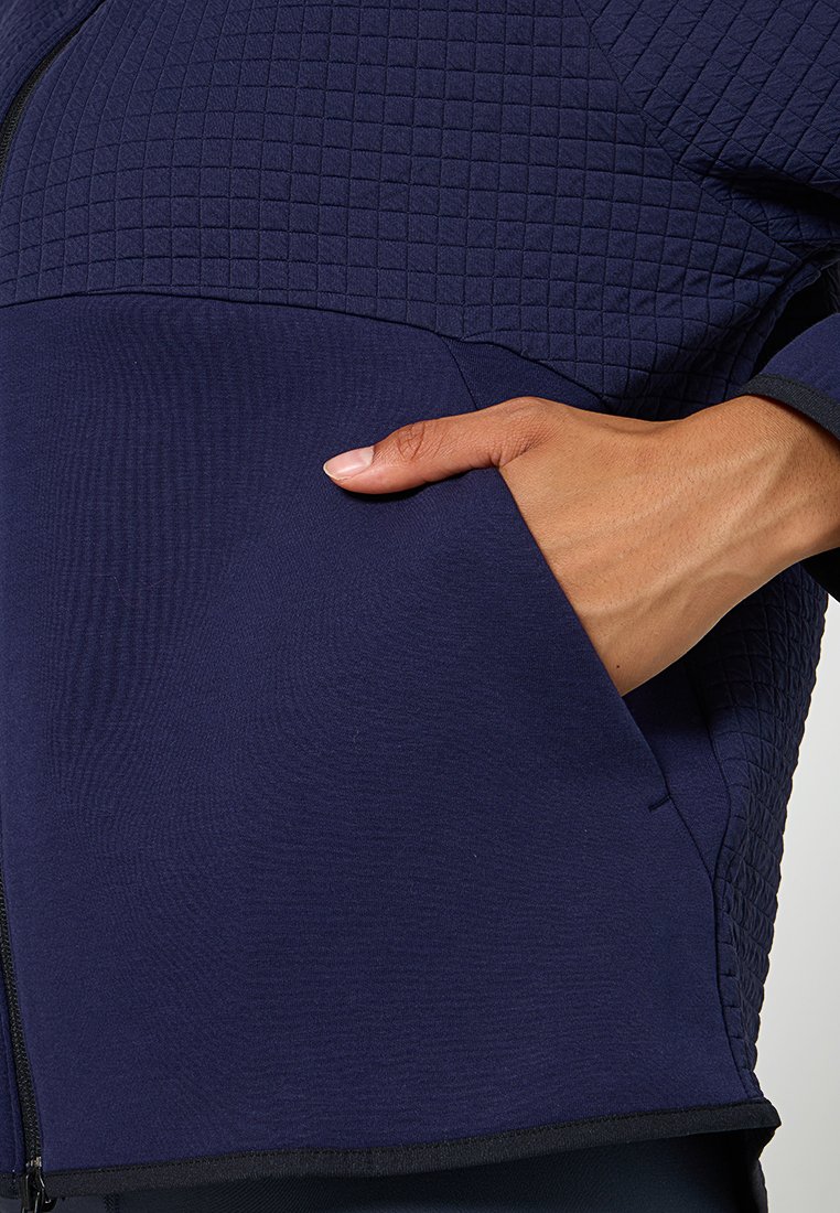 Chaqueta acolchada azul marino que presenta un panel de tela suave, cierre de cremallera y una mano en el bolsillo lateral, destacando texturas contrastantes.