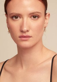 Pendientes de tono dorado con un diseño cilíndrico y un pequeño detalle de perla. El modelo lleva un maquillaje neutro y un top de tirantes negro.