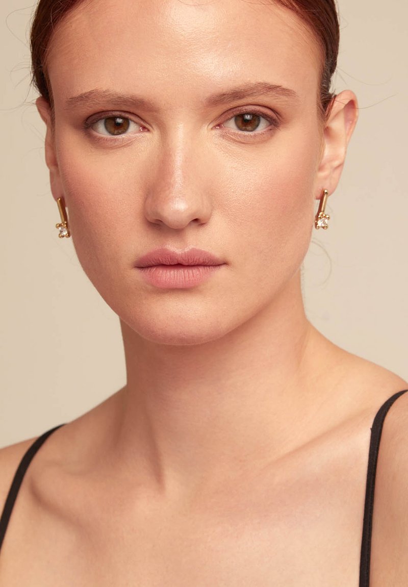 Pendientes de tono dorado con un diseño cilíndrico y un pequeño detalle de perla. El modelo lleva un maquillaje neutro y un top de tirantes negro.