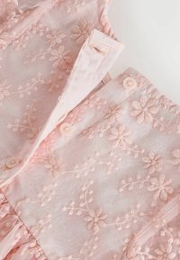 Tissu rose pâle transparent avec motifs floraux brodés et petits boutons, montrant un gros plan d'un fermoir délicat de vêtement et des coutures.