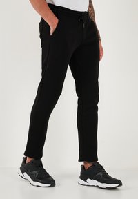 Schwarze Jogginghosen im Slim-Fit, mit Kordelzugbund, seitlichen Taschen und glattem Material. Kombiniert mit schwarzen Sneakers mit weißen Sohlen.