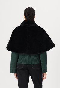 Svart cape i konstpäls med hög krage, bärs över en grön stickad tröja. Har en texturerad ribbad design och korta ärmar.