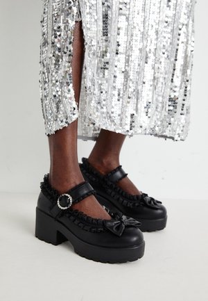 Zapatos Mary Jane negros con tacón grueso, adorno de volantes, detalle de lazo y hebilla con brillantina, combinados con una falda plateada de lentejuelas que llega hasta la mitad de la pantorrilla.