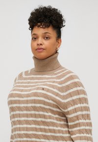 Pulover cu guler înalt, în dungi bej și crem, cu un model de tricotaj cu cabluri și accent cu logo pe piept. Material moale și ușor.