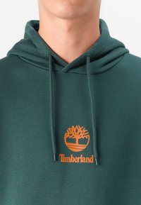 Felpa verde in cotone, con tasca frontale e cordini. Un logo arancione Timberland con un design ad albero è centrato sul petto.