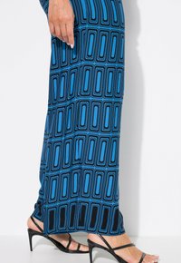 Modrá maxi šaty s černým geometrickým vzorem, vyrobené z měkké látky, v kombinaci s černými sandály na vysokém podpatku se zavazováním.