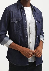 Camisa de manga larga de denim oscuro con un suéter gris claro debajo. Presenta dos bolsillos en el pecho, botones blancos y mangas arremangadas.