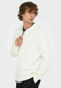 Weißer gerippter Pullover mit einem Reißverschlusskragen und langen Ärmeln, über einem schwarz-weißen gestreiften Hemd getragen, kombiniert mit dunklen Hosen.