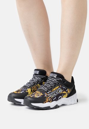 Zapatillas deportivas negras con patrones gráficos vibrantes en dorado y azul, con suela blanca texturizada y cordones negros. Marca en la lengüeta.