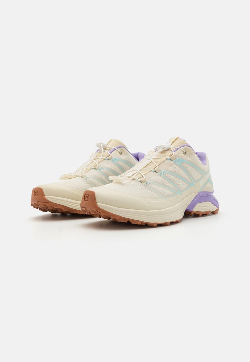 Zapatillas de running ligeras que cuentan con una parte superior de malla beige con acentos en azul y púrpura, una suela acolchada y una suela exterior texturizada en marrón.