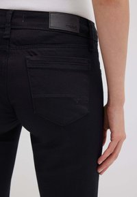Primo piano della parte bassa della schiena di una persona e della mano che indossa jeans neri con una tasca posteriore e un'etichetta in pelle "mavi" sul girovita.