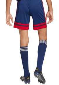 Marinefarbene Shorts mit roten und blauen Streifen, kombiniert mit knielangen marinefarbenen Socken, die drei weiße Streifen haben. Glänzende schwarze Fußballschuhe runden das Outfit ab.