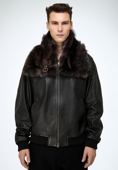 Noor mees, kes kannab musta nahktagi tumedapruuni karusnahkraega, seisab tavalise valge tausta ees.