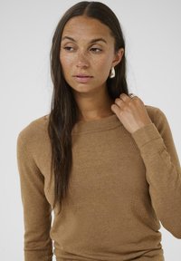 Maglione marrone a maniche lunghe con una texture morbida e scollatura a girocollo, indossato con uno stile informale. Il modello ha i capelli lunghi e indossa accessori.