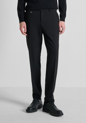 Hombre con pantalones de vestir negros de corte ajustado y zapatos de cuero negros, de pie contra un fondo liso de color gris claro.