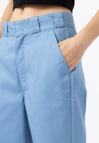 Person wearing high-waisted, light blue pants, with right hand partially inside front pocket, shown from midriff to thigh.