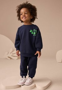 Granatowy sweatshirt i dopasowane spodnie wykonane z miękkiego materiału. Na swetrze znajduje się zielony nadruk traktora. Całość uzupełniają białe sneakersy.