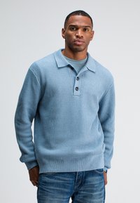 Helles blauer, gerippter Pullover mit langen Ärmeln, Kragen und einer Knopfleiste mit drei Knöpfen, kombiniert mit blauen Jeans. Strukturierter Stoff am Körper.