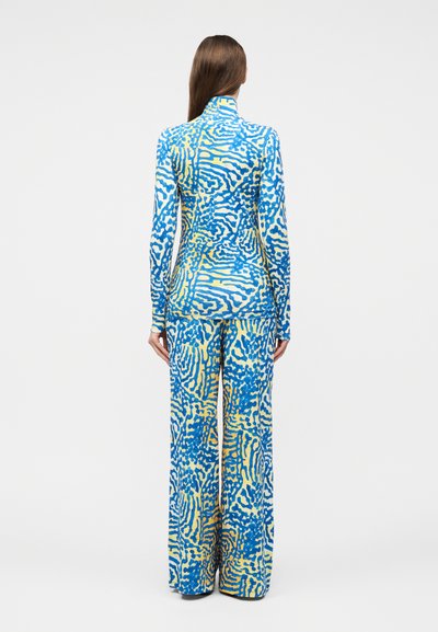 Γυναίκα που φαίνεται από πίσω, φοράει ταιριαστό μπλουζάκι με μακριά μανίκια και παντελόνι με φαρδιά πόδια, με αφηρημένο μοτίβο στροβιλιζόμενων μπλε και κίτρινων.