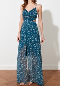 Femme portant une robe longue bleue à fleurs avec de fines bretelles, des découpes sur les côtés et une fente à l'avant, debout sur un sol en bois contre un mur blanc.