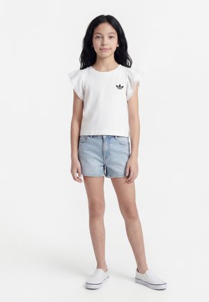 Κορίτσι με μακριά μαύρα μαλλιά, ντυμένη με λευκό πουκάμισο με βολάν στα μανίκια, ανοιχτό μπλε τζιν σορτς και λευκά παπούτσια slip-on, στέκεται σε λευκό φόντο.