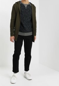 Mann trägt eine dunkelgrüne Kapuzenjacke mit Reißverschluss über einem grauen Rundhalsausschnittpullover, schwarze Hosen, weiße Socken und weiße Turnschuhe und steht in einem schlichten weißen Raum.