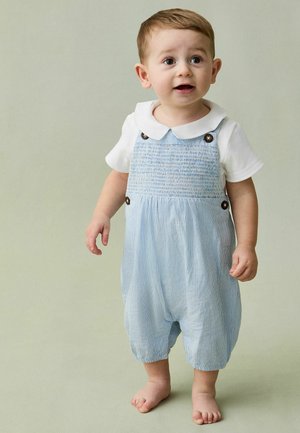 Mazs bērns stāv basām kājām, valkājot baltu kreklu un zilas svītrainas kombinezona bikses ar pogām, paskatoties nedaudz uz sāniem ar atvērtām mutēm.