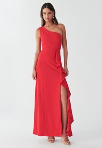 Vestido de noche rojo en tejido elástico, con diseño de un solo hombro, detalle de volante vertical y una abertura alta en la pierna. Textura suave.