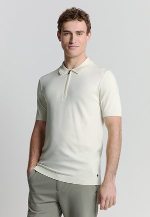Korte mouwen polo shirt in crème-kleurige brei. Heeft een ritskraag en een subtiele ribbeltextuur. Gecombineerd met lichtgroene broek.